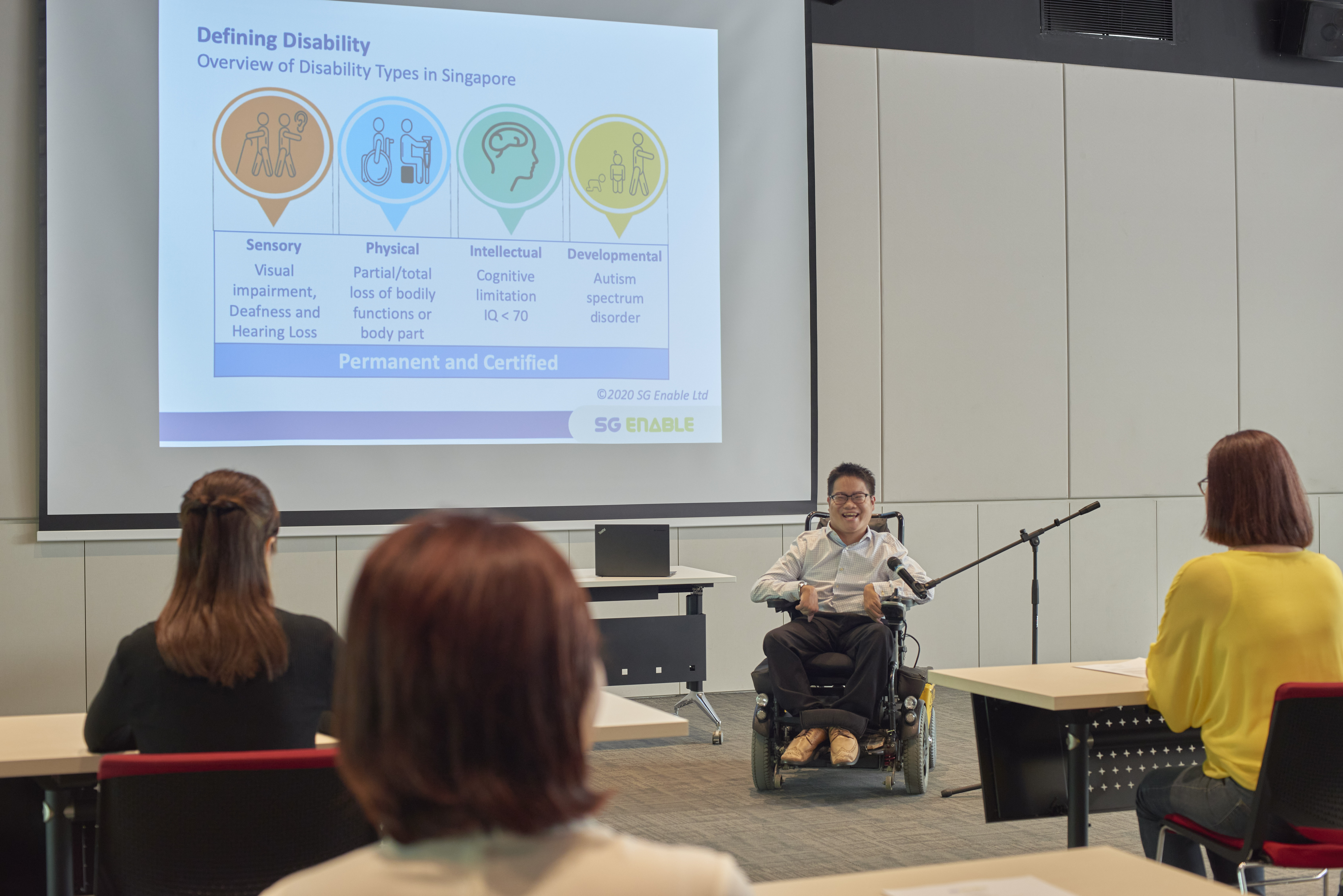 A man in a wheelchair happily giving a speech in a seminar room in front of a crowd.