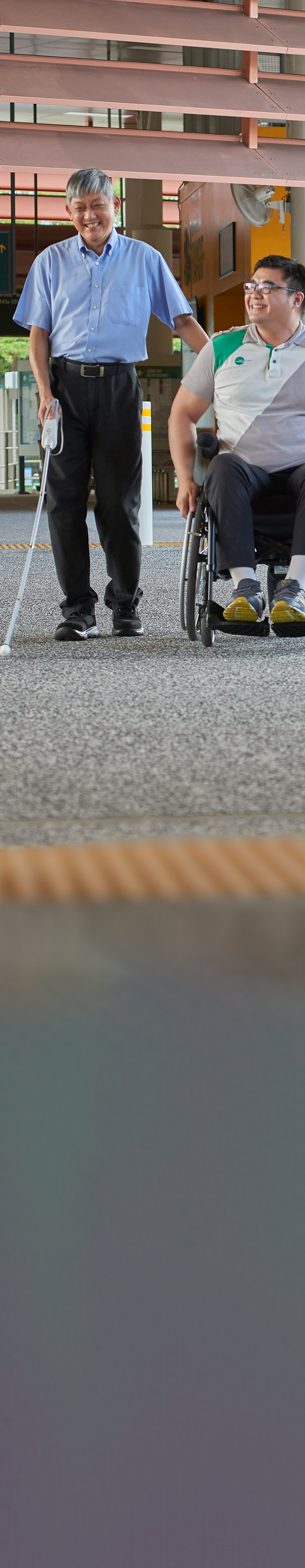 A man with visual impairment navigates his way around a bus interchange using the WeWALK Smart Cane. A guide in a wheelchair accompanies him as they chat and laugh.