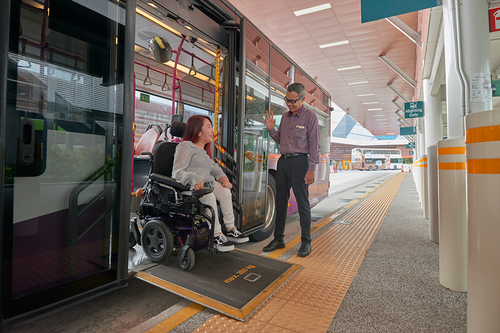 Bus captain standing outside a bus and smiling at a wheelchair user as she alights from the bus via a ramp.