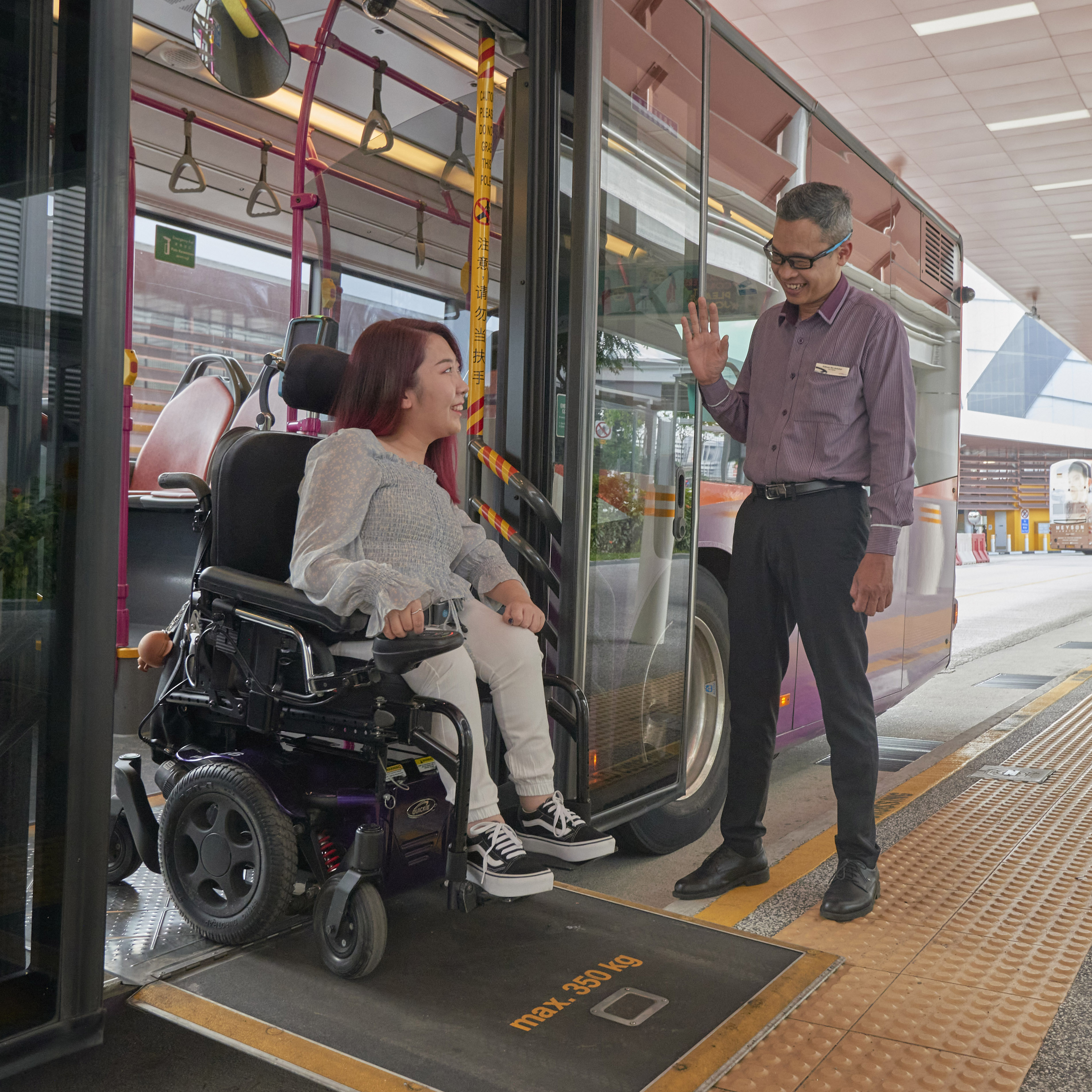 Bus captain standing outside a bus and smiling at a wheelchair user as she alights from the bus via a ramp.