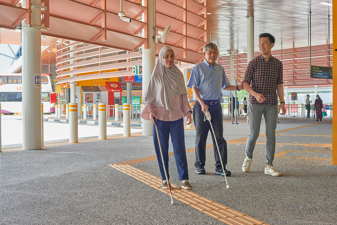 A man and woman, both with visual impairment, walking with another person without disability at a bus interchange.