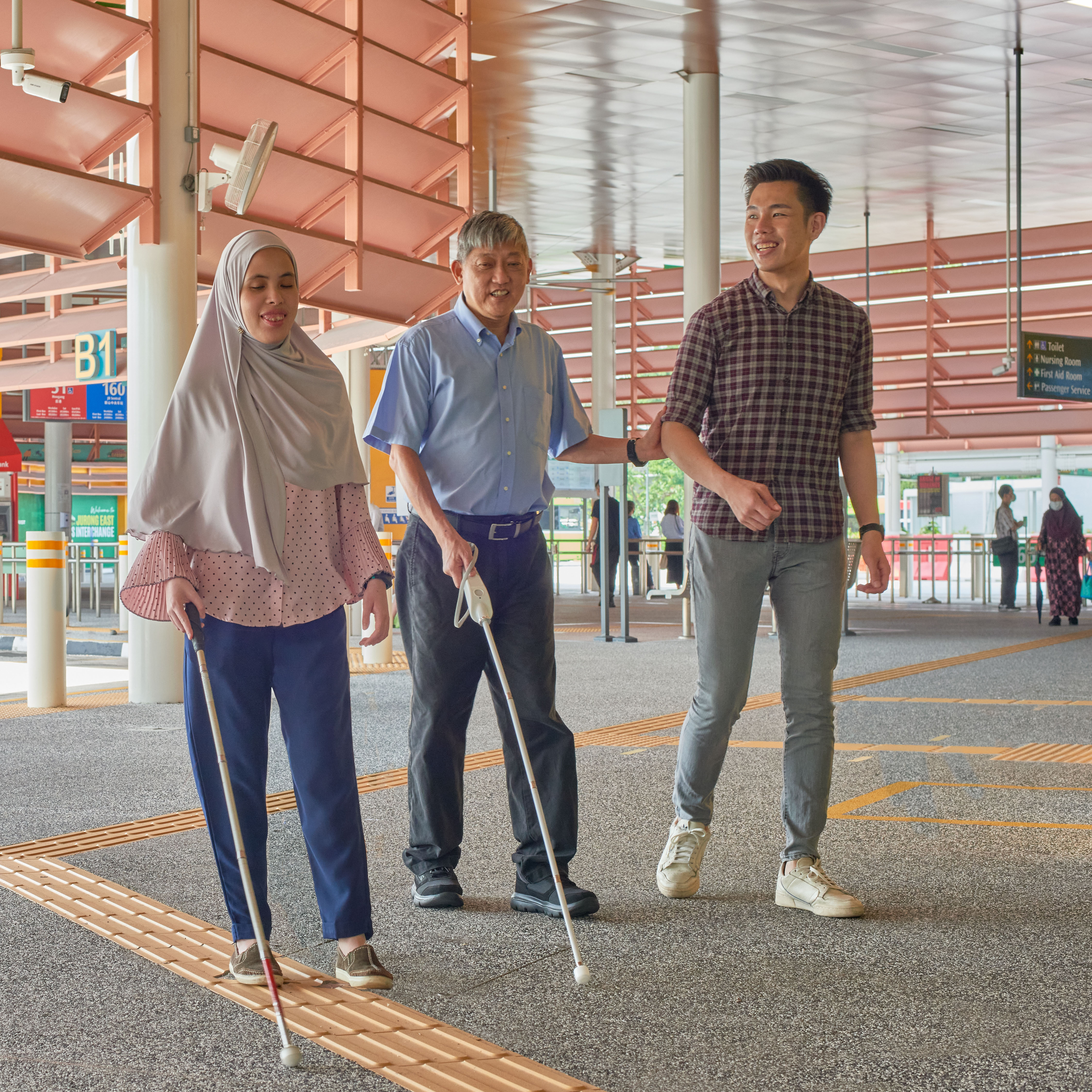 A man and woman, both with visual impairment, walking with another person without disability at a bus interchange.