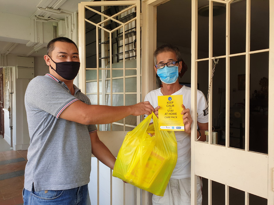 A masked SG Enable staff handing a masked resident a yellow care pack, with a pamphlet that reads "Keep Calm and Stay At Home Care Pack"
