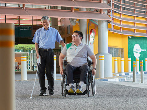 A smiling man with visual impairment walks with his cane and his hand on the shoulder of a wheelchair user, who is moving along with him at a bus interchange.