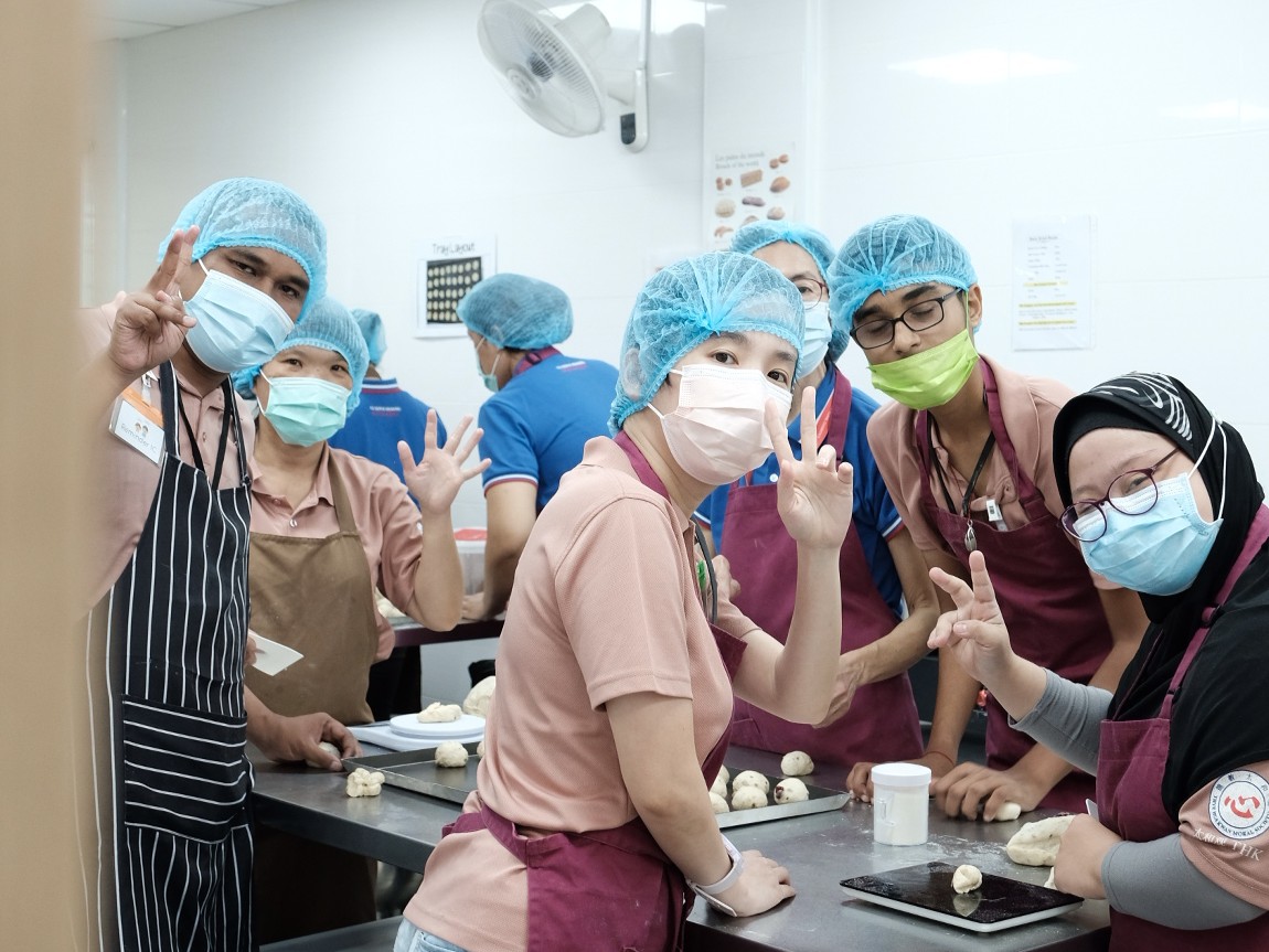 A group of persons with disabilities smiling and waving at the camera from a baking class.