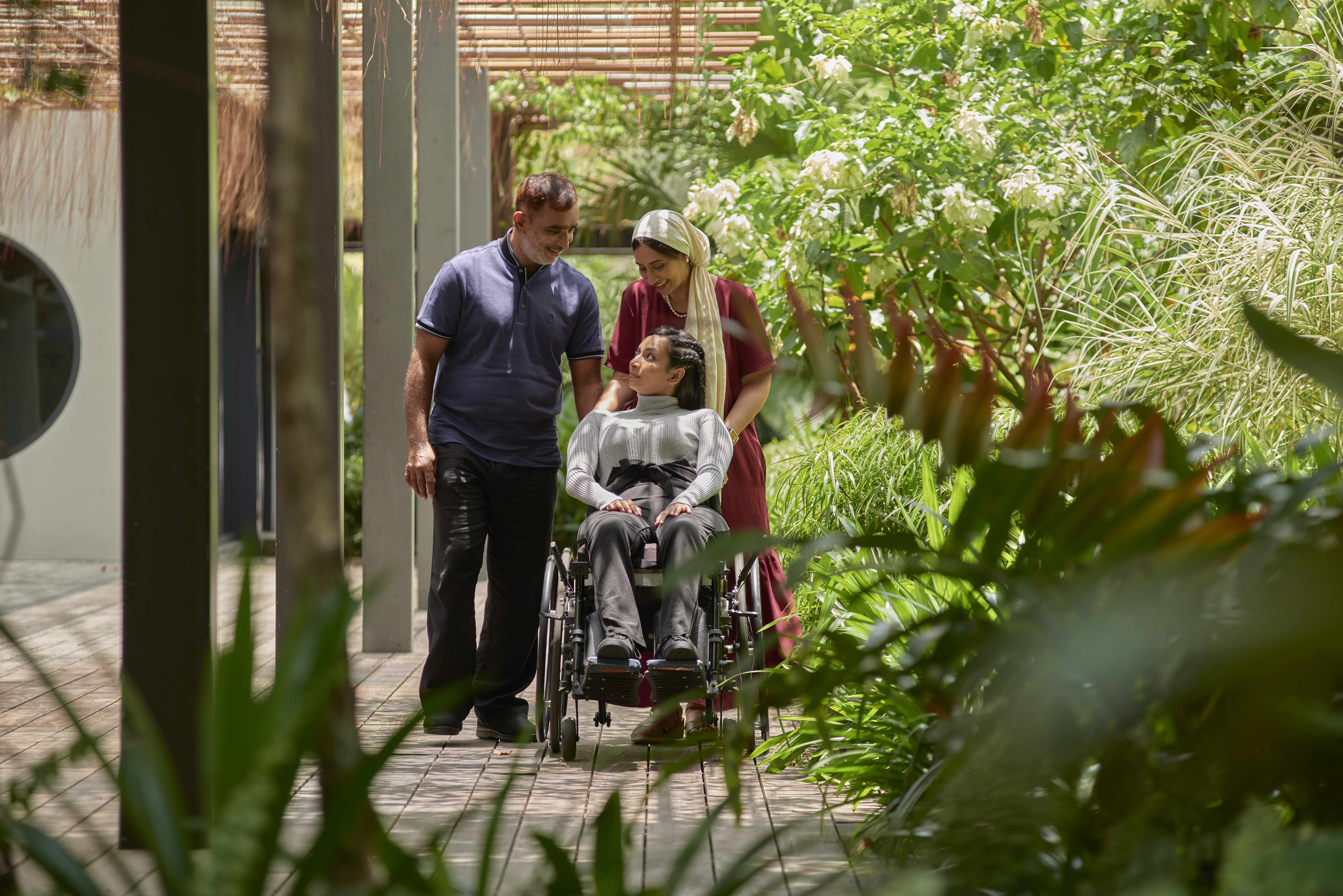 Surrounded by greenery, two caregivers and their care recipient on a wheelchair looks affectionately at one another