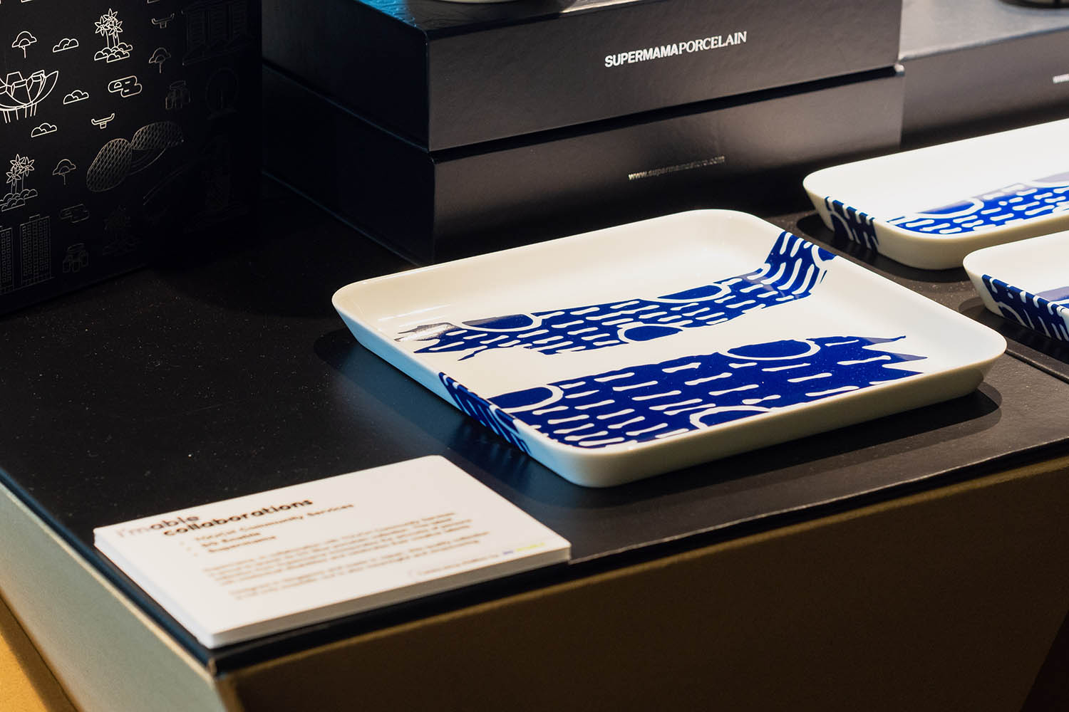 A display of a few porcelain plates with blue motifs, with black boxes in the background. The text on the boxes reads, SUPERMAMAPORCELAIN