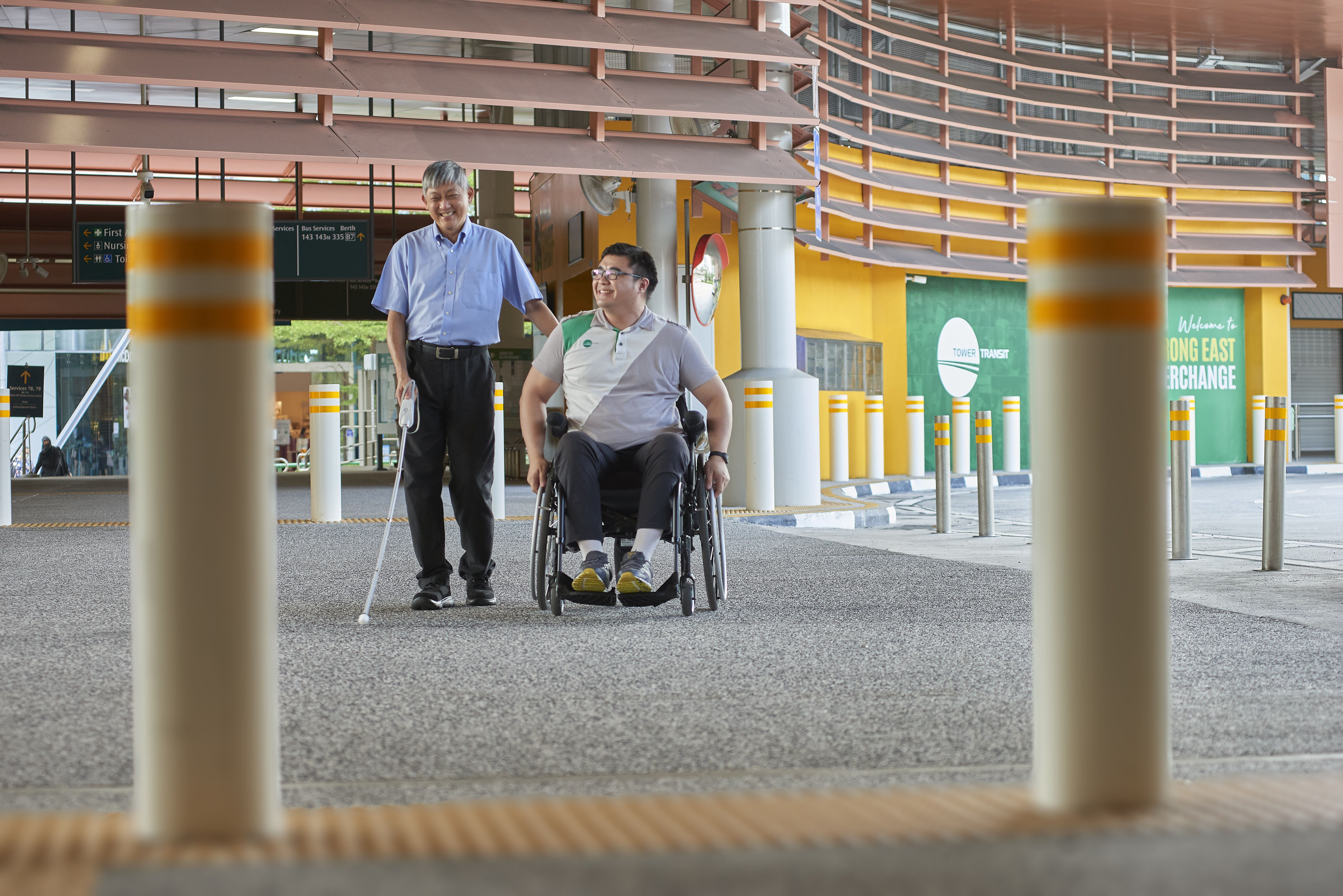 A wheelchair user guiding a man with a white cane along the bus interchange