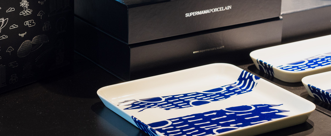 A display of a few porcelain plates with blue motifs, with black boxes in the background. The text on the boxes reads, SUPERMAMAPORCELAIN