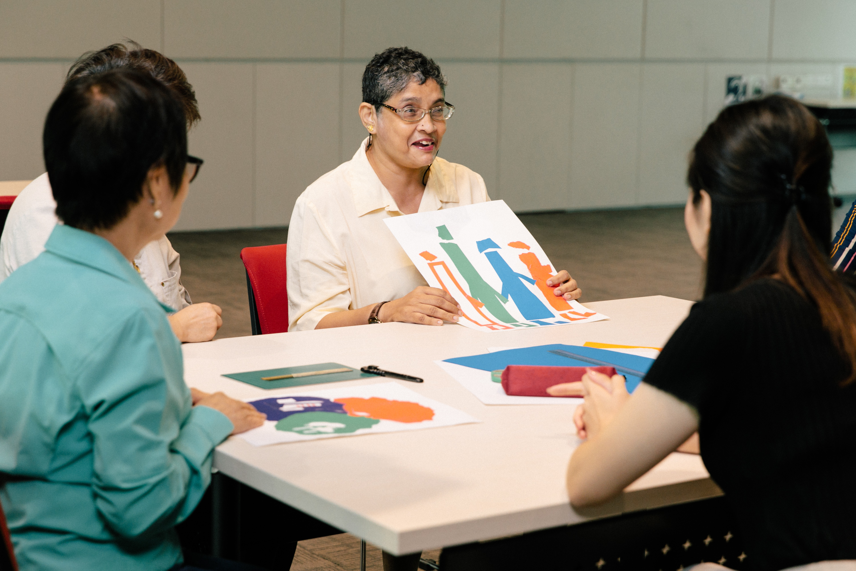 A short-haired lady holds up a piece of drawing as she talks to her companions.