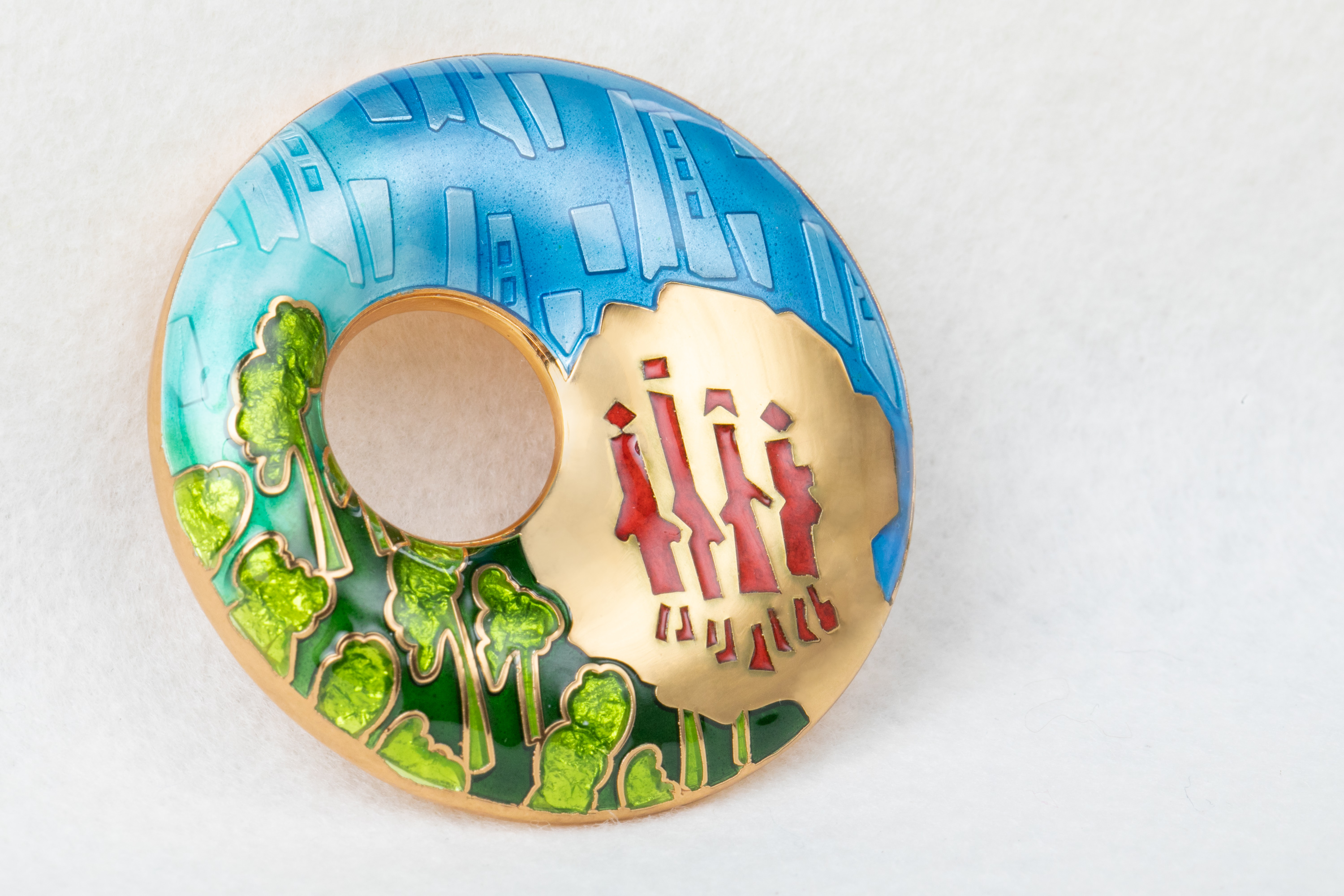 A round gold brooch with design of people, trees and buildings.
