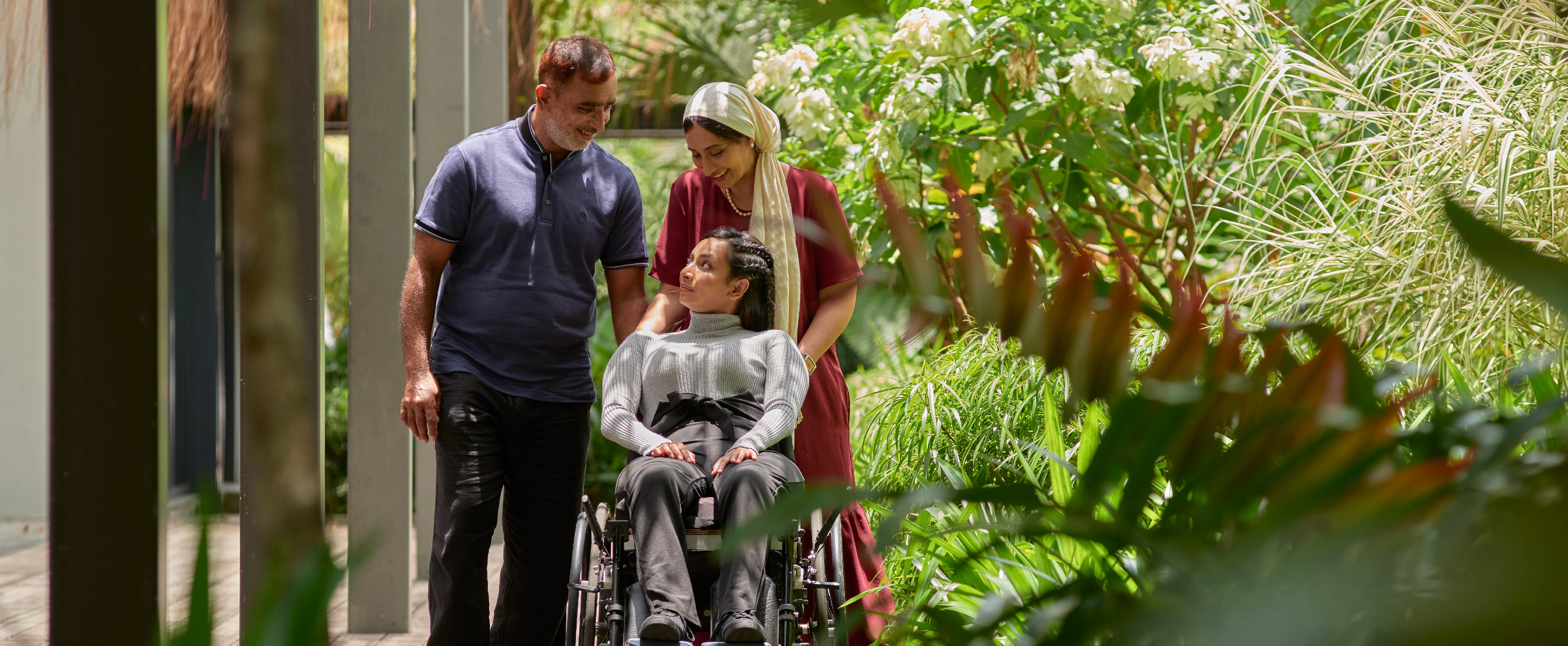 Surrounded by greenery, two caregivers and their care recipient on a wheelchair looks affectionately at one another
