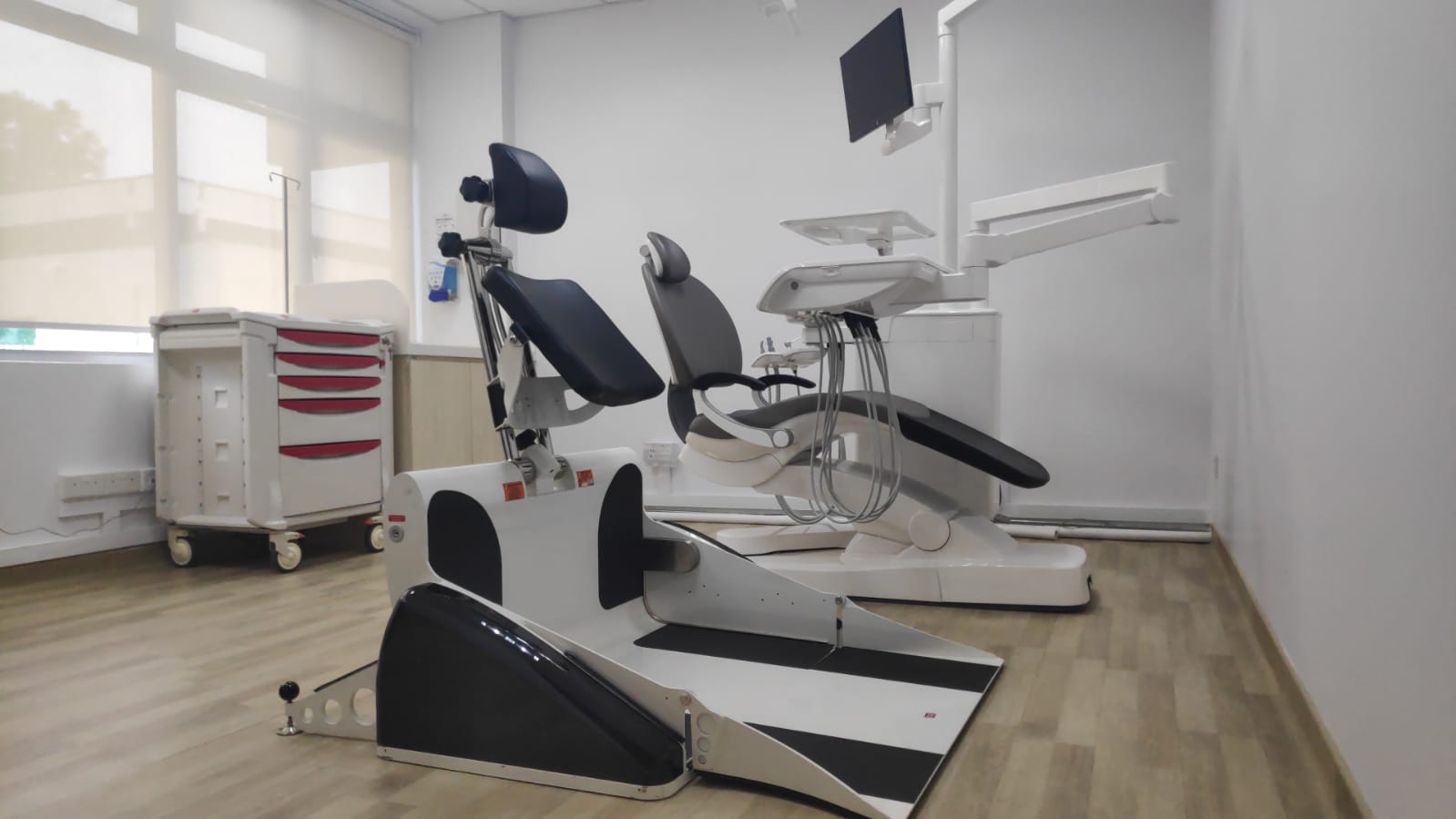 A wheelchair tilter positioned next to a regular dental chair in the dental clinic.