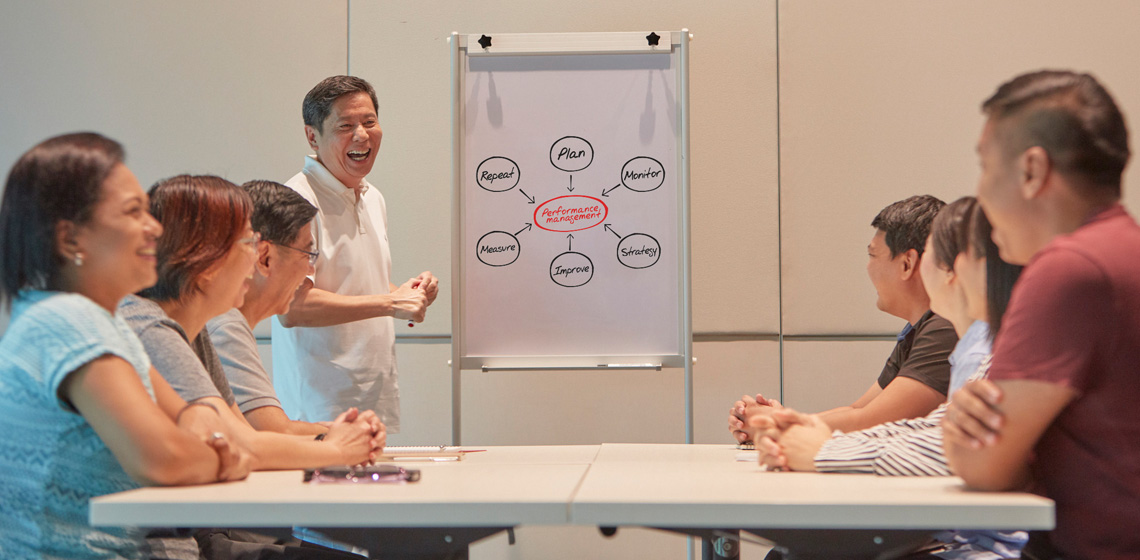 A group of people seated at two sides of a table, as another man stands in the centre and gives a presentation next to a whiteboard.