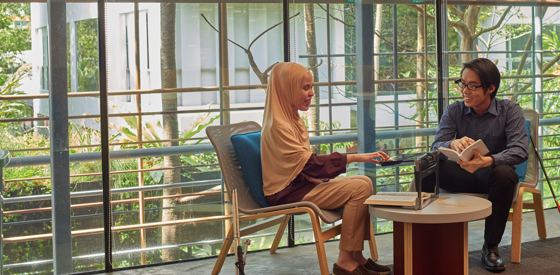 Two individuals seated at a coffee table having a conversation. One of them is using an electronic magnifier to read a book.