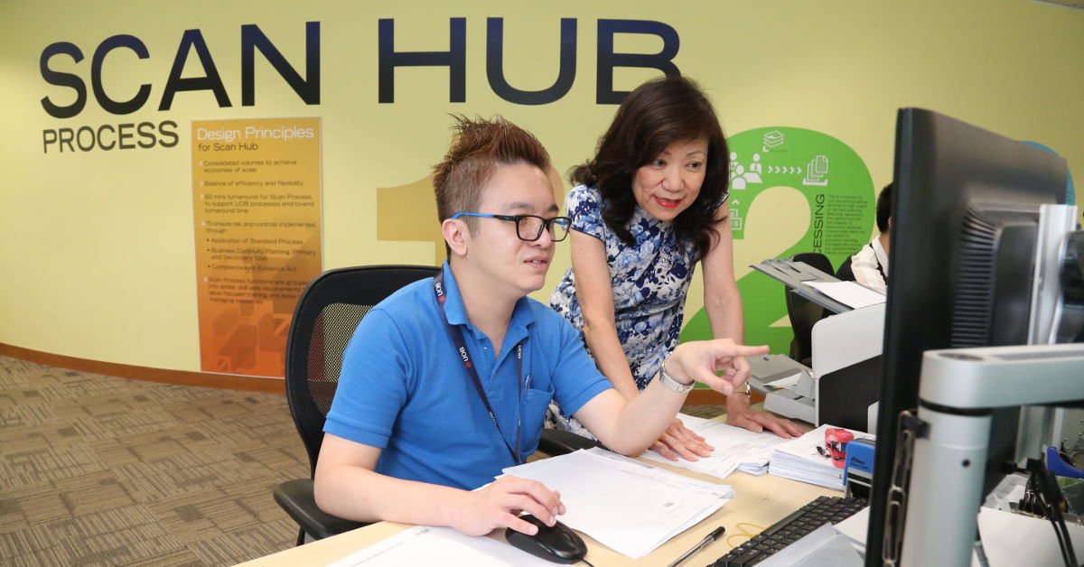 A female colleague gives some instructions to her male colleague in front of a computer.