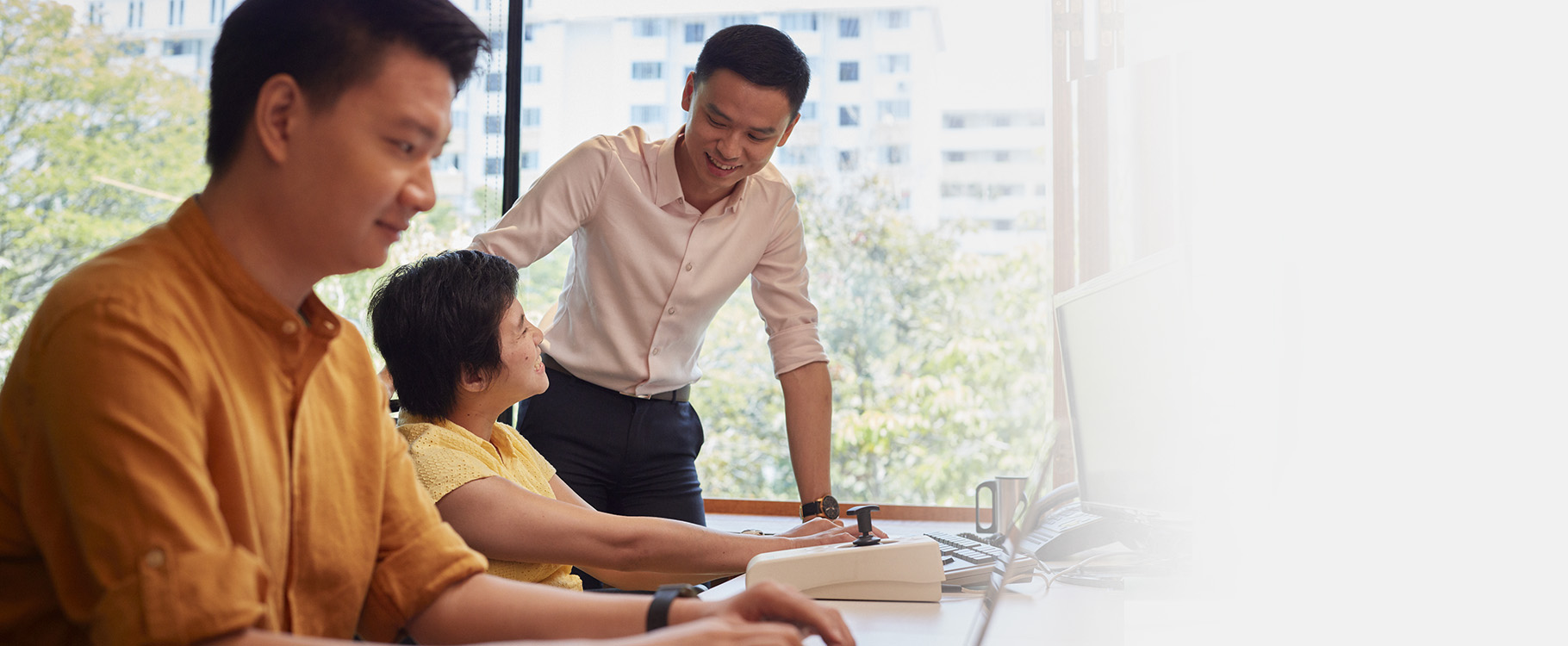 A man in the background is interacting with a woman with disability, while a man in the foreground works on his laptop.