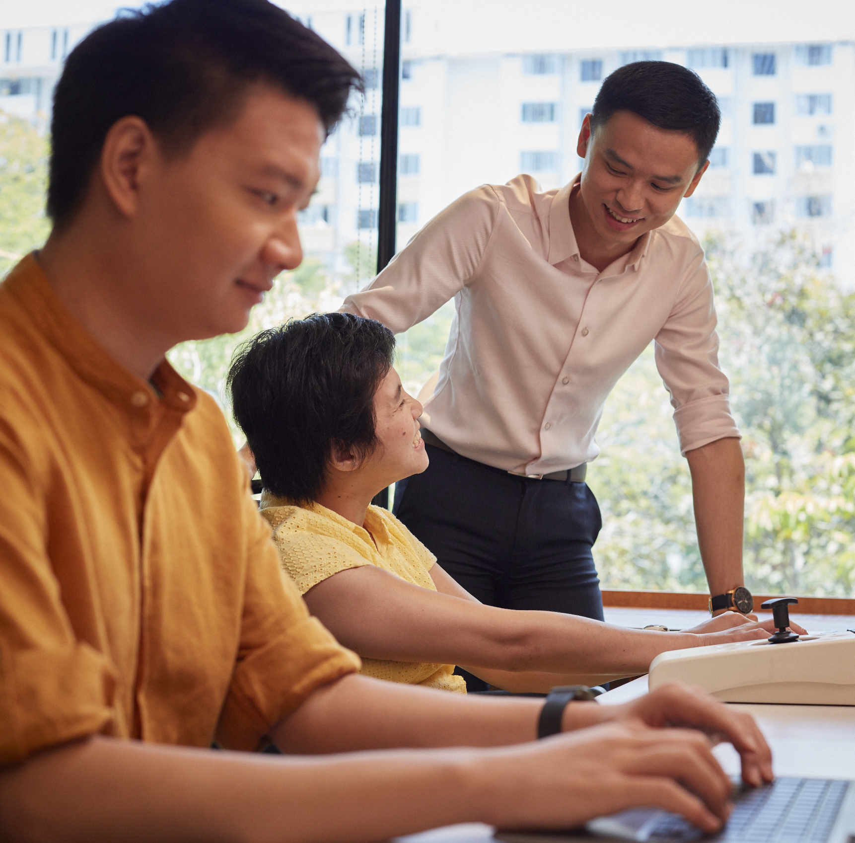 A man in the background is interacting with a woman with disability, while a man in the foreground works on his laptop.