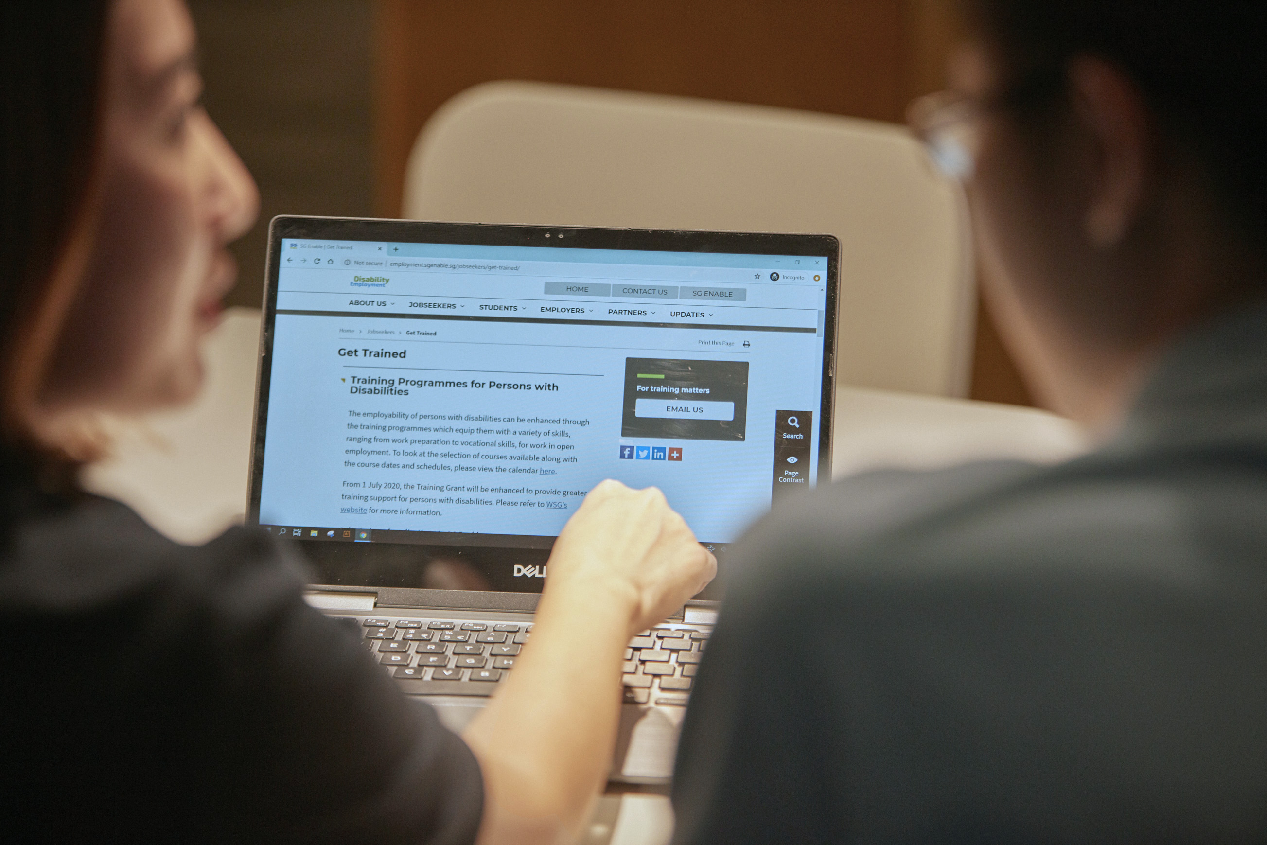 Two people in the foreground, with the man on the right looking at the laptop screen in the background, and the woman on the left looking to him as she uses the laptop.