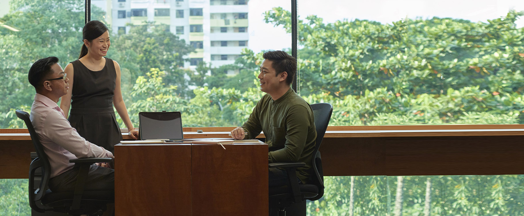 Three professionals, one on the right and seated across the other two, having a conversation at a work desk.