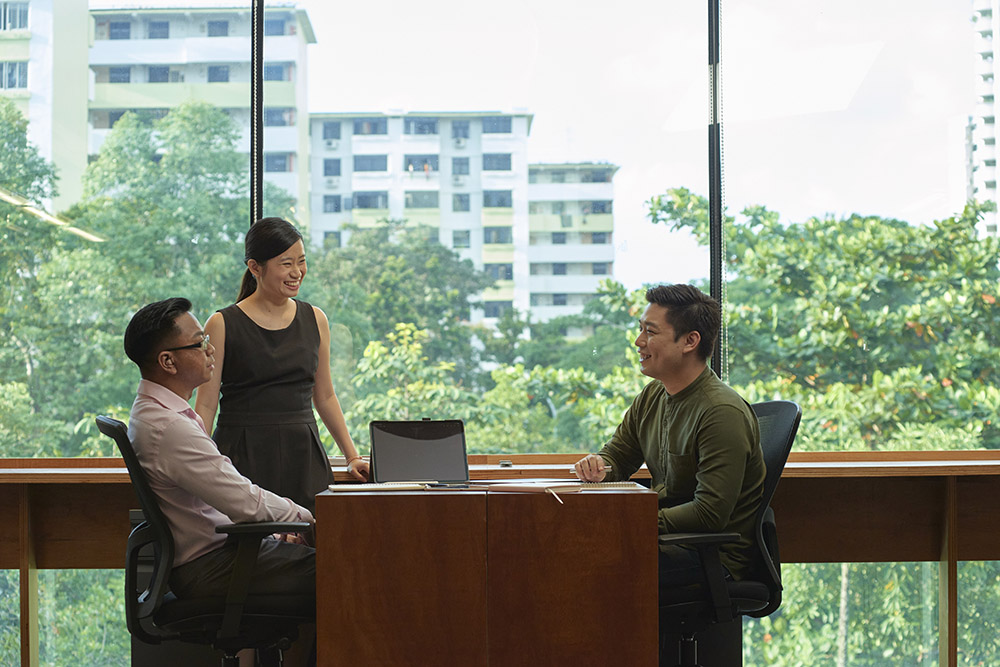 Three professionals, one on the right and seated across the other two, having a conversation at a work desk.Co