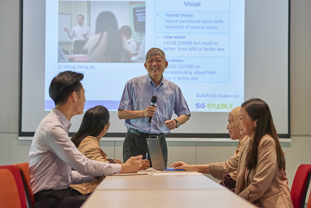 Four people seated at the same table and listening as a man with visual impairment gives a presentation.