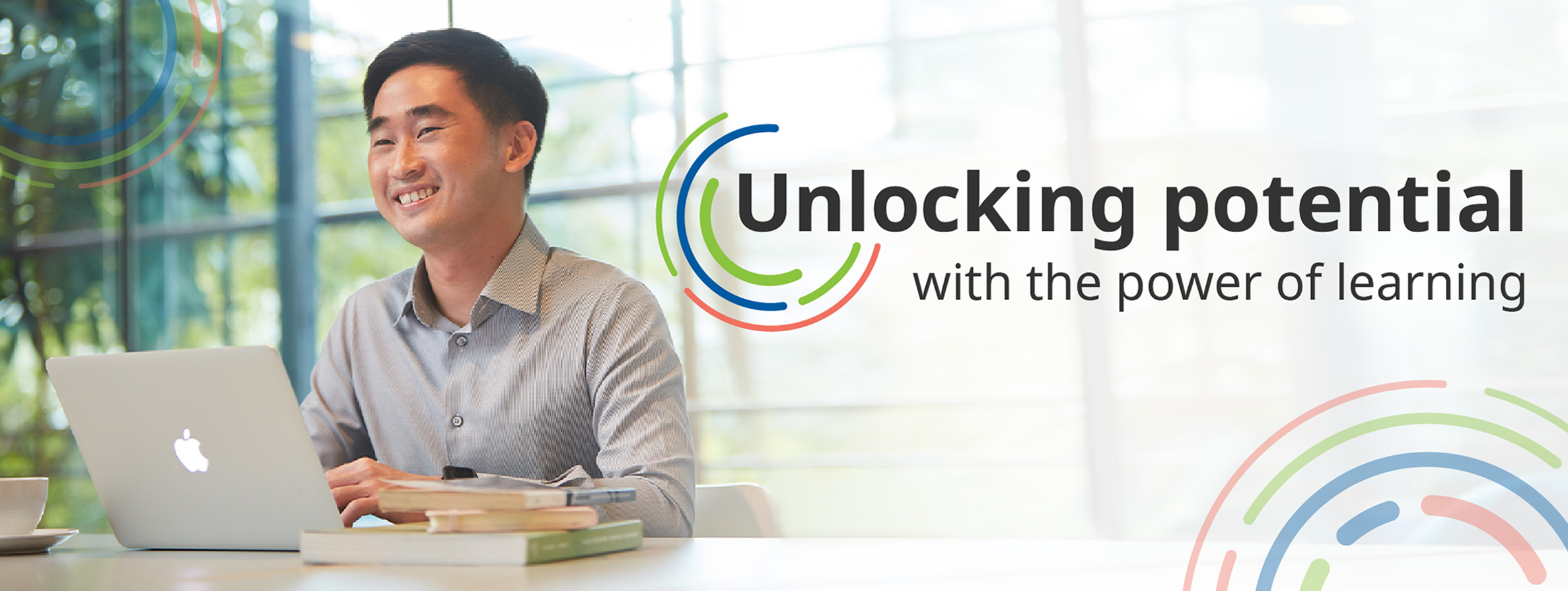 A man smiles as he works on his laptop. The text in the visual - which has illustrations of concentric rings in blue, green and coral orange - says "Unlocking potential with the power of learning".