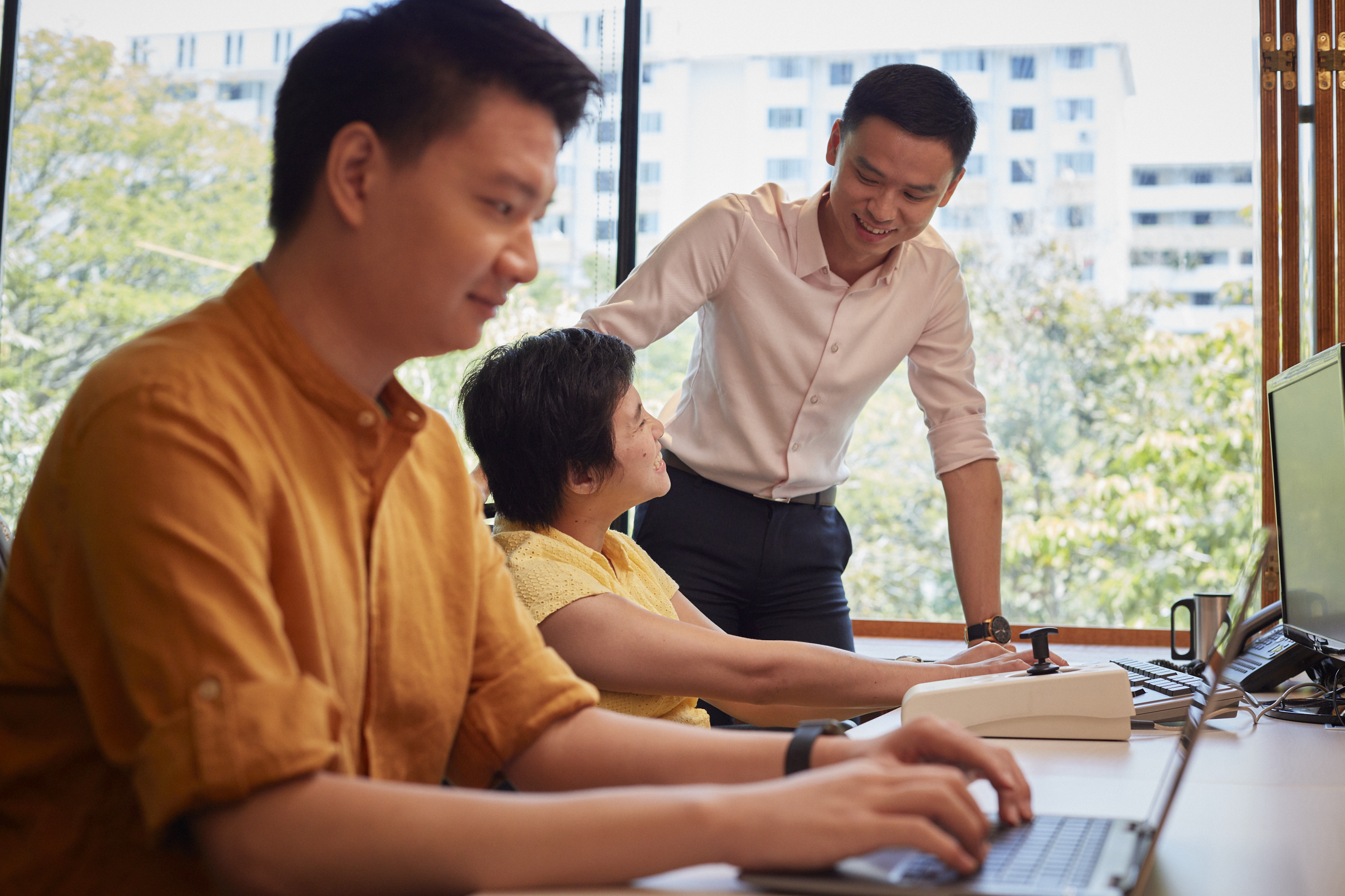 A man in the background is interacting with a woman with disability, while a man in the foreground works on his laptop.