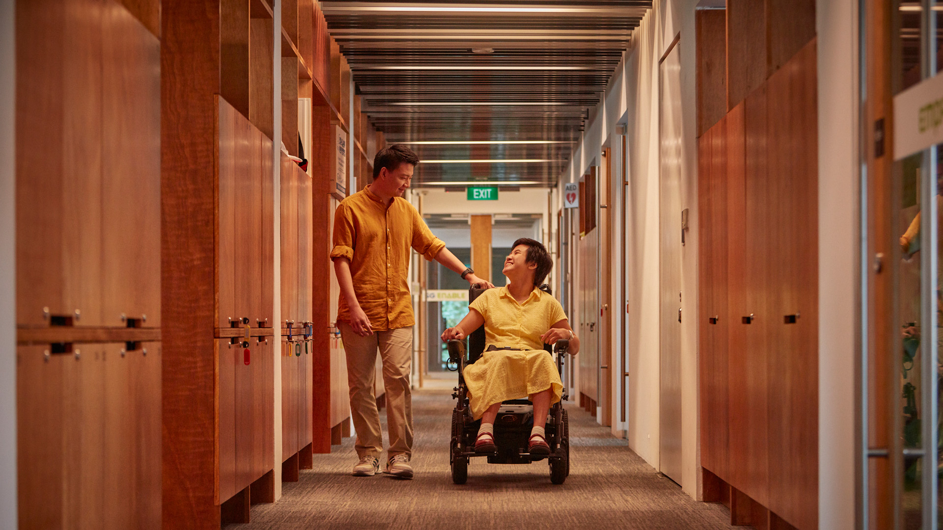 A woman in a wheelchair chats with a male colleague, as they move across an office corridor.