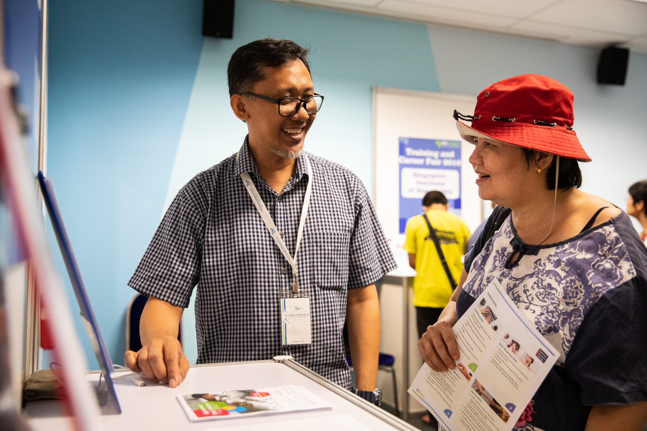 A bespectacled man speaks to a woman wearing a red bucket hat who is holding onto a brochure.