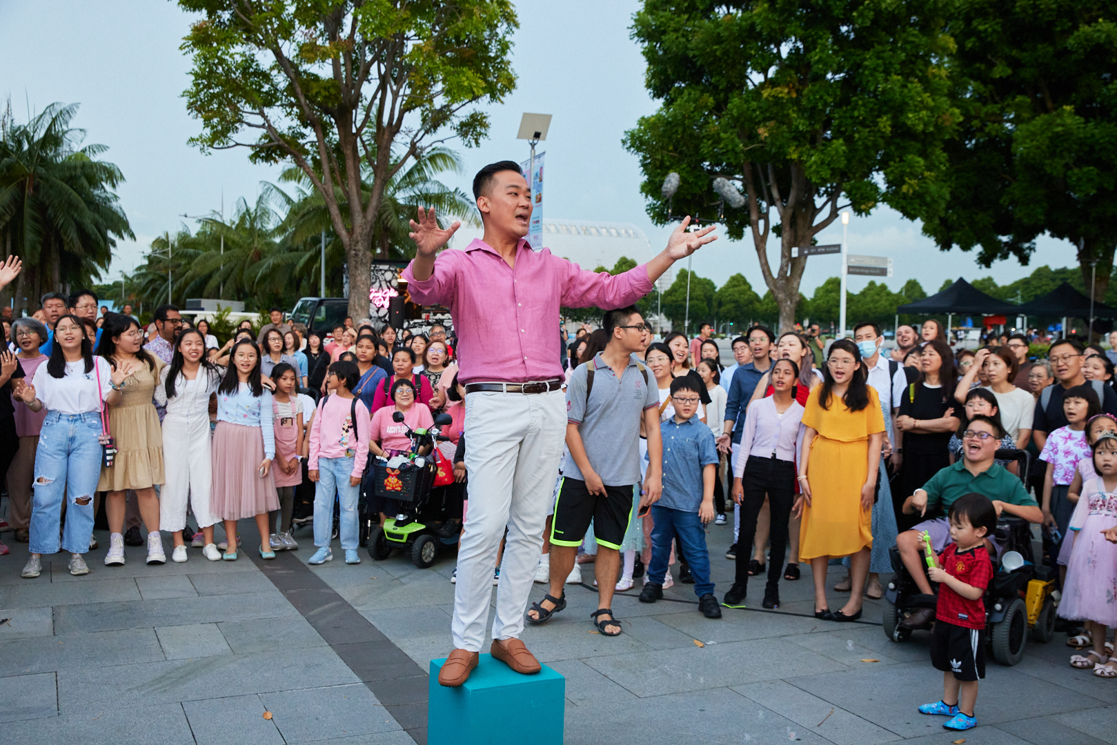 A man in pink stands on a blue box conducting a crowd in a rendition of 