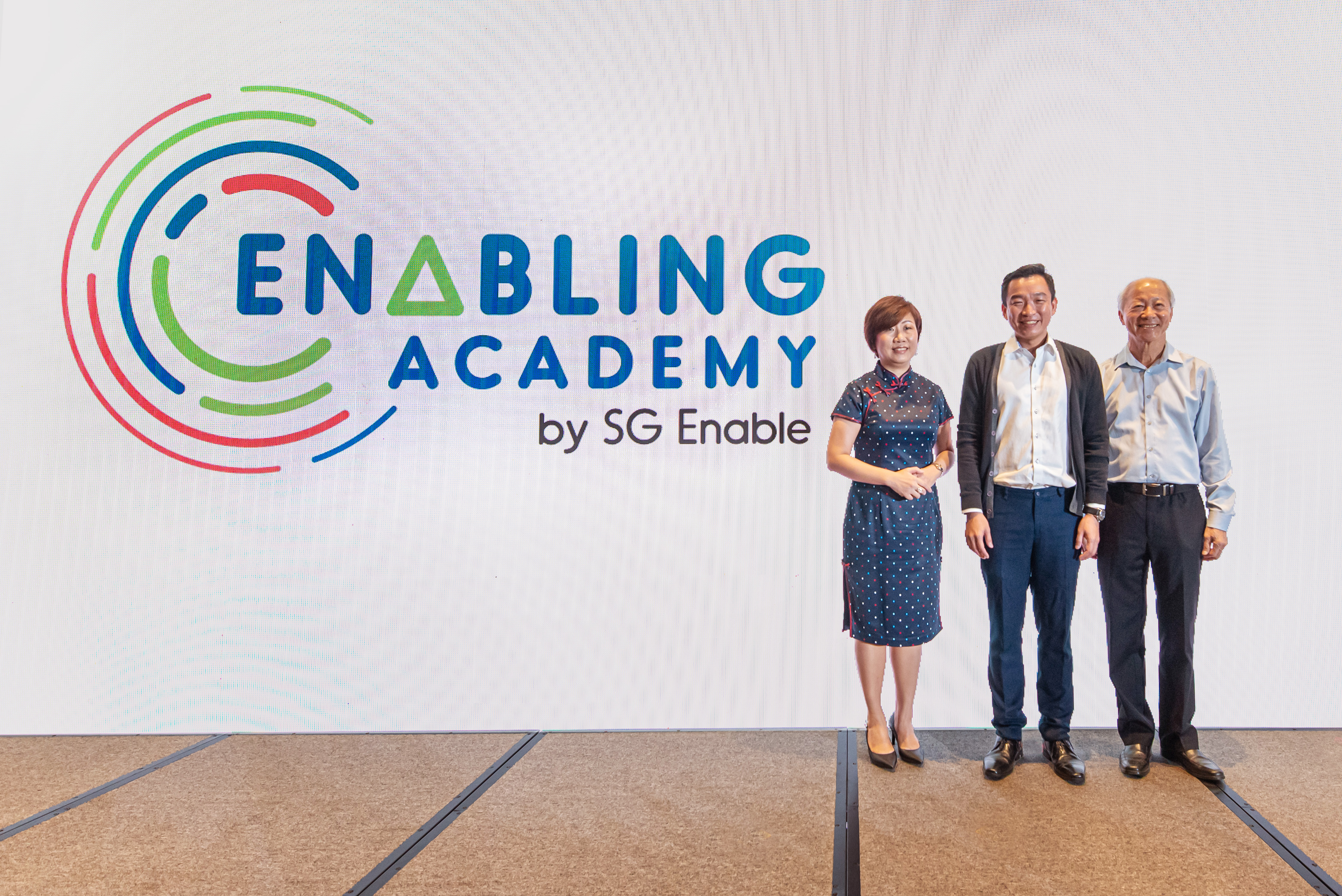 SG Enable’s CEO, Ms Ku Geok Boon, Parliamentary Secretary Mr Eric Chua and SG Enable’s Chairman Mr Moses Lee standing against a screen that displays Enabling Academy’s logo.