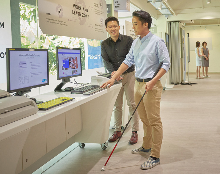 A man is seen guiding a visually impaired man to a braille device at Tech Able.