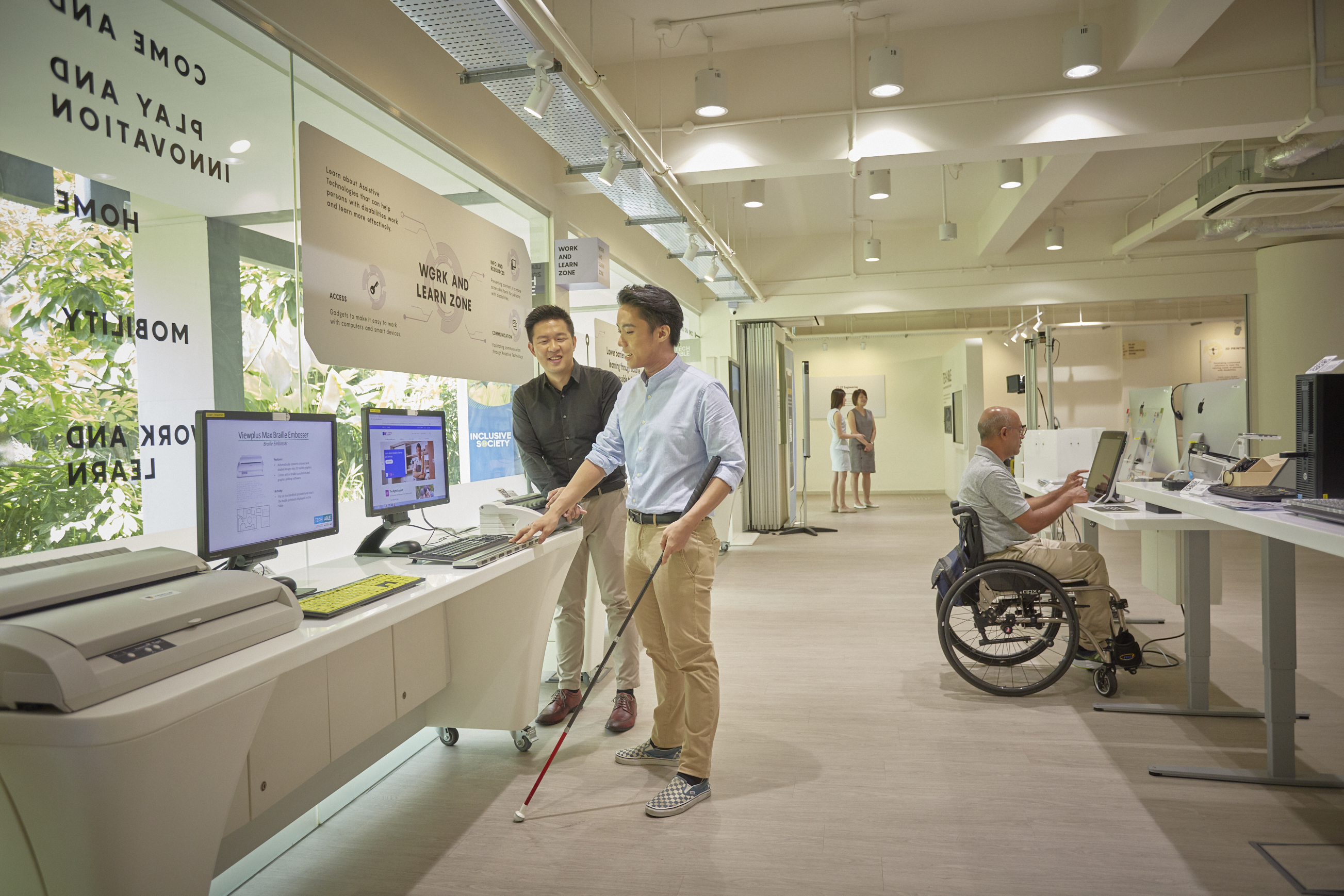 A person with visual impairment navigates Tech Able with his white cane, while behind him, a wheelchair user explores an AT device on display.